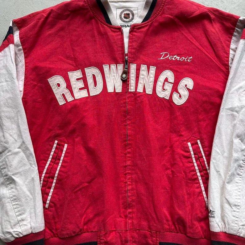 May include: Red and white baseball jacket with the words "Detroit Redwings" on the front. The jacket has white sleeves and a black zipper.