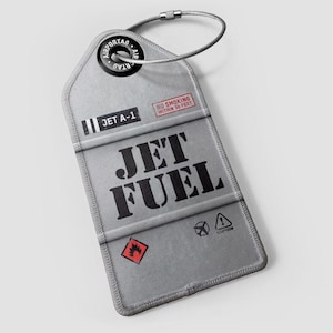 Puede incluir: Una etiqueta de equipaje gris con las palabras "JET FUEL" en letras negras grandes. La etiqueta también presenta el texto "JET A-1" y "NO SMOKING WITHIN 50 FEET". Es visible un símbolo de peligro de llama rojo y negro. La etiqueta está sujeta a un lazo de metal.
