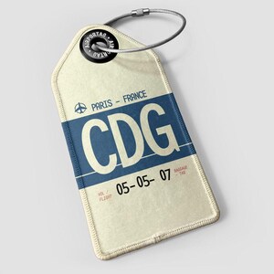 Puede incluir: Etiqueta de equipaje color crema con una franja azul y las letras "CDG". La etiqueta dice "Paris - France" y "05-05-07". Un lazo y un alambre metálicos aseguran la etiqueta. La etiqueta tiene un borde cosido.