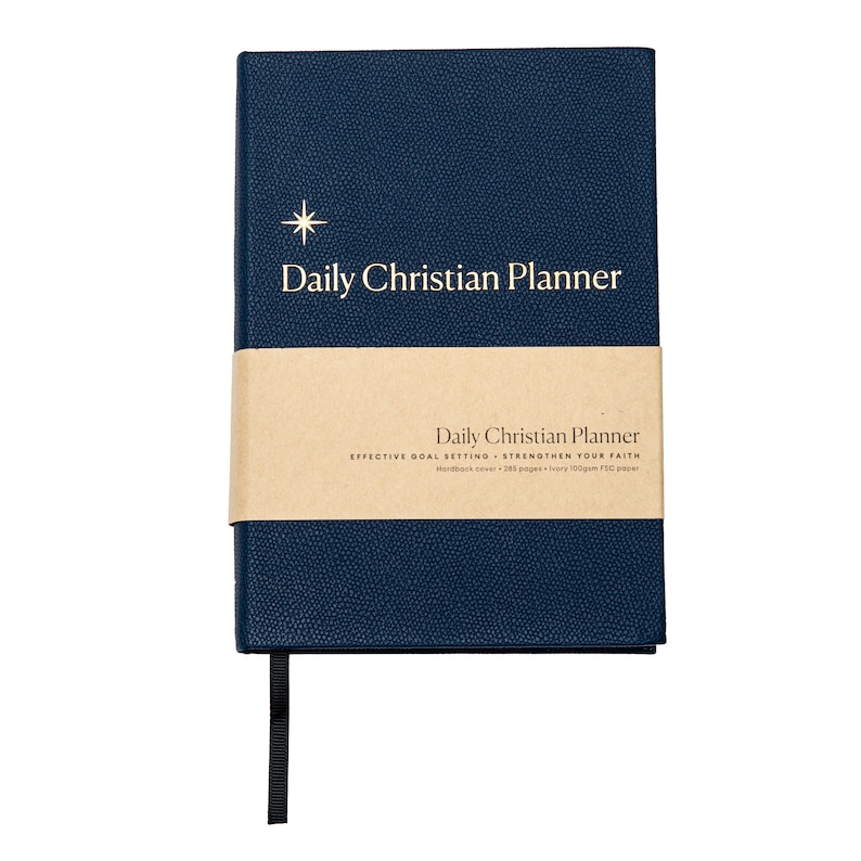 May include: A navy blue hardcover journal with a gold star and the text "Daily Christian Planner" on the front cover. The journal has a brown paper band with the text "Daily Christian Planner, Effective Goal Setting - Strengthen Your Faith, Hardback cover - 285 pages - Ivory 100gsm paper".