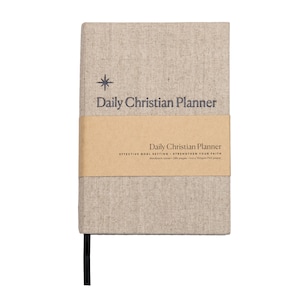 May include: A beige linen-covered journal with a brown paper band. The journal has a star and the text "Daily Christian Planner" printed on the front cover. The band has the text "Daily Christian Planner" and "Effective Goal Setting - Strengthen Your Faith" printed on it.