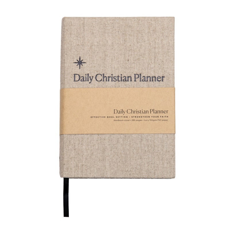 May include: A beige linen-covered journal with a brown paper band. The journal has a star and the text "Daily Christian Planner" printed on the front cover. The band has the text "Daily Christian Planner" and "Effective Goal Setting - Strengthen Your Faith" printed on it.