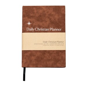 May include: A brown faux leather daily Christian planner with a gold star and the text "Daily Christian Planner" on the front cover. The spine is black and the planner has a brown paper band with the text "Daily Christian Planner" and "Effective Goal Setting - Strengthen Your Faith" on it.