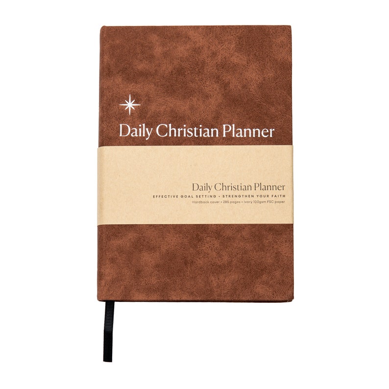May include: A brown faux leather daily Christian planner with a gold star and the text "Daily Christian Planner" on the front cover. The spine is black and the planner has a brown paper band with the text "Daily Christian Planner" and "Effective Goal Setting - Strengthen Your Faith" on it.