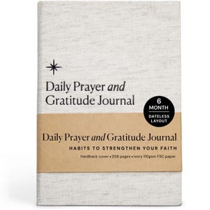 May include: A hardcover journal with the text "Daily Prayer and Gratitude Journal" in black. The journal has a light beige linen cover and a brown band with additional text. A black circle indicates a 6-month dateless layout.