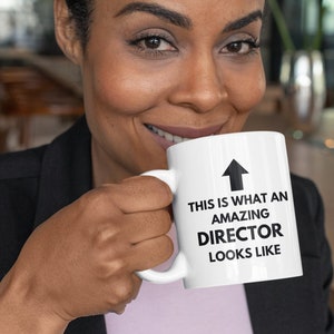 May include: A white ceramic mug with the text "THIS IS WHAT AN AMAZING DIRECTOR LOOKS LIKE" and an upward-pointing arrow. The mug is held by a person wearing a black blazer.