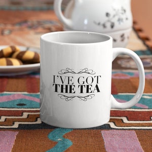 May include: White ceramic mug with the text "I'VE GOT THE TEA" in black, with a white teapot and a plate of cookies in the background. The mug has a curved handle and sits on a colorful patterned tablecloth.