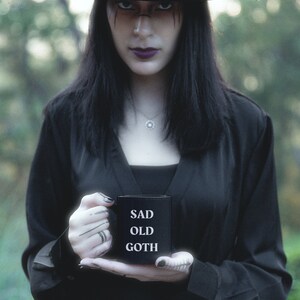 May include: A person wearing a black hat and black clothing holds a black mug with the words "SAD OLD GOTH" in white. The person has dark makeup and a silver necklace. The background is blurred green.