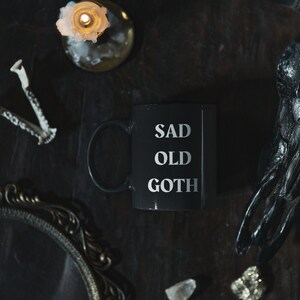 May include: A black mug with the words "SAD OLD GOTH" in silver lettering. The mug is on a dark surface with a lit candle, decorative mirror, crystals, and a raven skull. The overall theme is gothic.