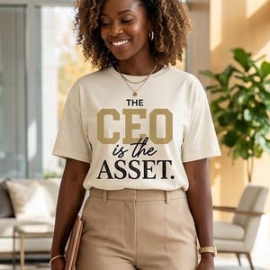 May include: A cream-colored short-sleeve t-shirt featuring the text THE CEO is the ASSET in gold and black lettering, paired with tan high-waisted trousers, white sneakers, and a brown leather handbag. The outfit includes a gold necklace and watch.
