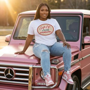 May include: A white t-shirt with the text "Soft Life Strong Boundaries" in an oval design. The shirt is paired with light blue ripped jeans and pink sneakers. The person is sitting on the hood of a pink SUV.