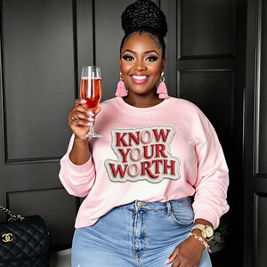 Può includere: Una felpa rosa chiaro con la scritta "KNOW YOUR WORTH" in rosso e bianco. La persona indossa jeans blu e tiene in mano un bicchiere di champagne rosa. La persona indossa anche orecchini a nappa rosa e un orologio d'oro.