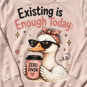 May include: A light pink sweatshirt featuring a graphic of a duck wearing black sunglasses and a polka-dot headscarf. The duck holds a pink coffee cup labeled ZERO GIVEN with a heart. Text above reads Existing is Enough Today: with stars.