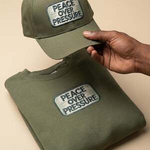 May include: Olive green baseball cap and sweatshirt set. Both items feature a rectangular patch with the text "PEACE OVER PRESSURE" in a stylized font. The cap is held above the sweatshirt, showcasing the matching design. The background is a solid, neutral color.