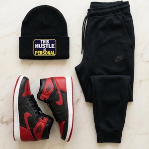Puede incluir: Un gorro negro con el texto "THIS HUSTLE IS PERSONAL" está junto a zapatillas altas negras y rojas y un pantalón de chándal negro. Las zapatillas tienen un swoosh y detalles en rojo. El pantalón de chándal tiene un logotipo de Nike.