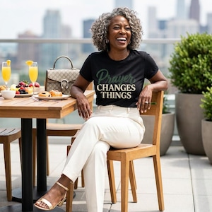 Peut inclure: Un t-shirt noir à manches courtes avec l'inscription "Prayer CHANGES THINGS" en lettres vertes et crème. Le t-shirt est associé à un pantalon couleur crème et à des sandales dorées, formant une tenue décontractée et coordonnée.