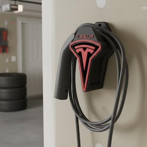 May include: Black Tesla electric vehicle charger mounted on a beige wall. The charger has a red Tesla logo and a coiled charging cable. The garage setting includes tyres and tools.