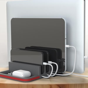 May include: A gray charging station with a red accent, holding four black phones and a white wireless charging pad. The station is sitting on a wooden table next to a laptop and a white cable.