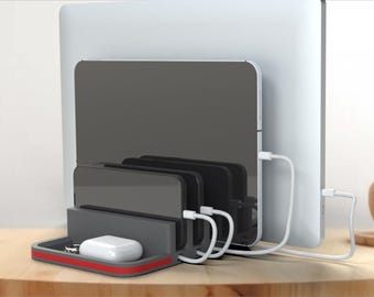 May include: A gray charging station with a red accent, holding four black phones and a white wireless charging pad. The station is sitting on a wooden table next to a laptop and a white cable.
