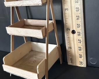 May include: A three-tier wooden shelf unit with a natural wood finish. The shelf unit is approximately 25 cm tall and is made of thin wooden slats. The shelf unit is ideal for displaying small items or plants.