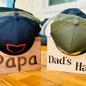 May include: Two wooden hat racks with the words "Papa" and "Dad's Hats" engraved on them. The racks are holding baseball caps, one rack has three black, green, and blue caps, the other rack has two green and black caps.