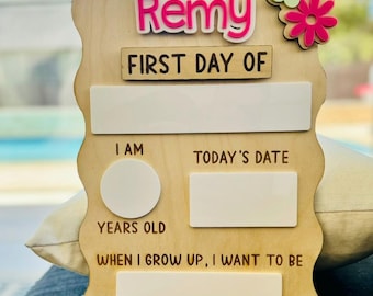 May include: A wooden sign with a wavy edge, decorated with pink and green flowers. The sign says "Remy First Day Of" and has blank spaces for writing in the child's age, today's date, and what they want to be when they grow up.