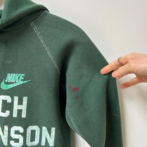K&ouml;nnte beinhalten: Dunkelgr&uuml;ner Nike-Hoodie mit dem Text "Coach Johnson" in wei&szlig;en Blockbuchstaben. Das Nike-Logo ist gr&uuml;n. Rote Flecken befinden sich auf dem rechten &Auml;rmel. Der Hoodie hat eine Kapuze und eine Vordertasche.