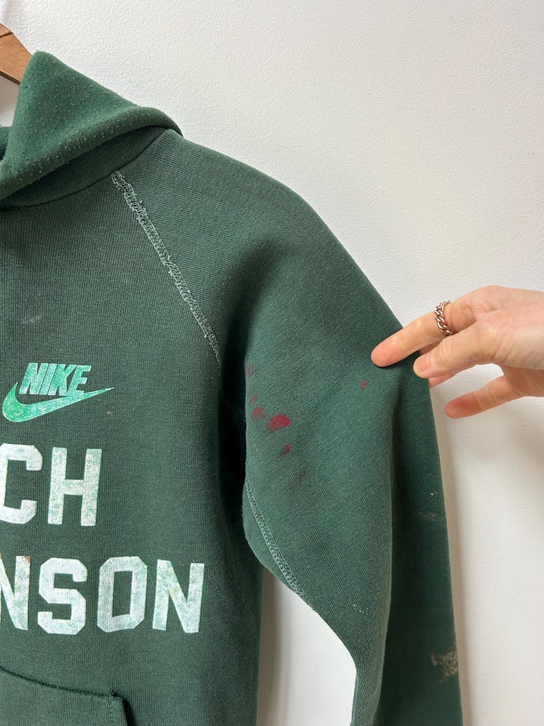 K&ouml;nnte beinhalten: Dunkelgr&uuml;ner Nike-Hoodie mit dem Text "Coach Johnson" in wei&szlig;en Blockbuchstaben. Das Nike-Logo ist gr&uuml;n. Rote Flecken befinden sich auf dem rechten &Auml;rmel. Der Hoodie hat eine Kapuze und eine Vordertasche.