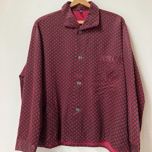 May include: A button-down shirt with a red and black diamond pattern. The shirt has a collar, a chest pocket, and three visible buttons. The shirt is hanging on a wooden hanger.