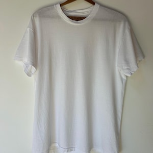 May include: A plain white short-sleeved t-shirt with a crew neckline. The shirt is displayed on a wooden hanger. Inside the collar, a small brown tag is visible with the word CLEANERS printed in black text. The garment has a simple design.