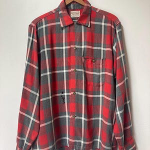 Peut inclure: Une chemise à manches longues boutonnée avec un motif à carreaux rouge, gris et blanc. La chemise présente un col pointu, une poche poitrine unique avec fermeture à bouton et une série de boutons sur le devant. Usure visible et petits trous.