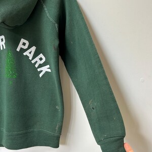 K&ouml;nnte beinhalten: Dunkelgr&uuml;ner Kapuzenpullover mit den W&ouml;rtern "PARK" und "R" in wei&szlig;en Blockbuchstaben. Zwischen den Buchstaben befindet sich eine gr&uuml;ne Baumgrafik. Der Pullover hat gerippte B&uuml;ndchen und Gebrauchsspuren.