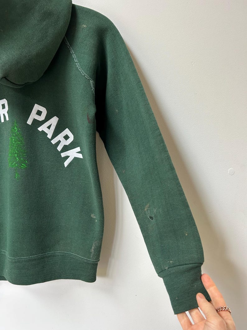 K&ouml;nnte beinhalten: Dunkelgr&uuml;ner Kapuzenpullover mit den W&ouml;rtern "PARK" und "R" in wei&szlig;en Blockbuchstaben. Zwischen den Buchstaben befindet sich eine gr&uuml;ne Baumgrafik. Der Pullover hat gerippte B&uuml;ndchen und Gebrauchsspuren.