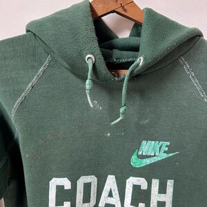 K&ouml;nnte beinhalten: Ein dunkelgr&uuml;ner Nike-Hoodie mit dem Wort "COACH" und dem Nike-Logo in Wei&szlig; und Gr&uuml;n. Der Hoodie hat eine Kapuze mit Kordelzug und h&auml;ngt an einem Holzb&uuml;gel.