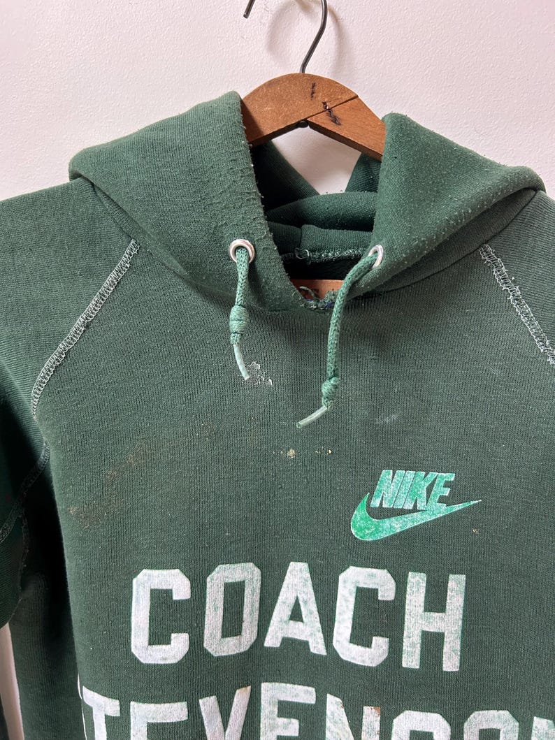 K&ouml;nnte beinhalten: Ein dunkelgr&uuml;ner Nike-Hoodie mit dem Wort "COACH" und dem Nike-Logo in Wei&szlig; und Gr&uuml;n. Der Hoodie hat eine Kapuze mit Kordelzug und h&auml;ngt an einem Holzb&uuml;gel.