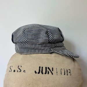 Può includere: Un berretto da macchinista a righe hickory blu navy e bianco con una visiera corta e rigida. Il berretto poggia su una borsa di tela beige con il testo nero stampato a stencil S.S. JUNIOR. Il cappello ha una corona arrotondata e una fascia.