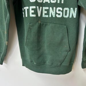 K&ouml;nnte beinhalten: Ein dunkelgr&uuml;ner Kapuzenpullover mit dem Text "COACH STEVENSON" in wei&szlig;en Blockbuchstaben. Das Nike-Logo ist &uuml;ber dem Text zu sehen. Der Pullover hat eine Vordertasche und weist Gebrauchsspuren auf.
