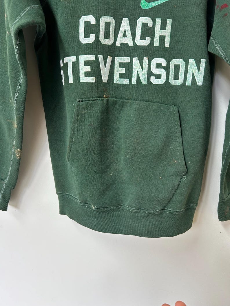 K&ouml;nnte beinhalten: Ein dunkelgr&uuml;ner Kapuzenpullover mit dem Text "COACH STEVENSON" in wei&szlig;en Blockbuchstaben. Das Nike-Logo ist &uuml;ber dem Text zu sehen. Der Pullover hat eine Vordertasche und weist Gebrauchsspuren auf.