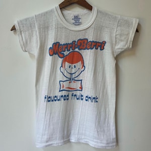 May include: A white short-sleeve t-shirt featuring a graphic of a cartoon character drinking from a pouch. The text reads Merri-Berri and flavoured fruit drink in blue and orange. The shirt is displayed on a wooden hanger against a plain wall.