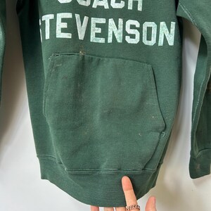 K&ouml;nnte beinhalten: Dunkelgr&uuml;nes Sweatshirt mit dem wei&szlig;en Aufdruck "COACH TEVENSON". Das Nike-Logo ist &uuml;ber dem Text sichtbar. Das Sweatshirt hat eine Vordertasche und einen gerippten Saum.