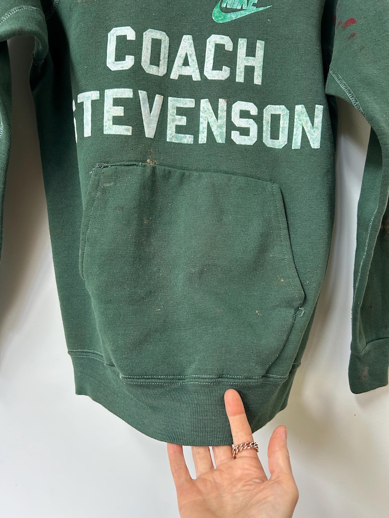 K&ouml;nnte beinhalten: Dunkelgr&uuml;nes Sweatshirt mit dem wei&szlig;en Aufdruck "COACH TEVENSON". Das Nike-Logo ist &uuml;ber dem Text sichtbar. Das Sweatshirt hat eine Vordertasche und einen gerippten Saum.