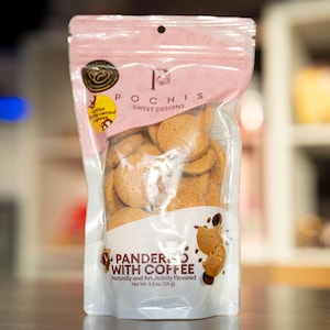 May include: A pink resealable bag of Pochis Sweet Designs Pandered with Coffee cookies. The bag contains a variety of round, light brown cookies. The bag is labeled with the text "Pochis Sweet Designs" and "Pandered with Coffee".