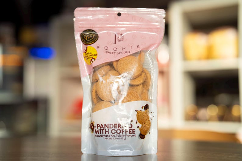 May include: A pink resealable bag of Pochis Sweet Designs Pandered with Coffee cookies. The bag contains a variety of round, light brown cookies. The bag is labeled with the text "Pochis Sweet Designs" and "Pandered with Coffee".