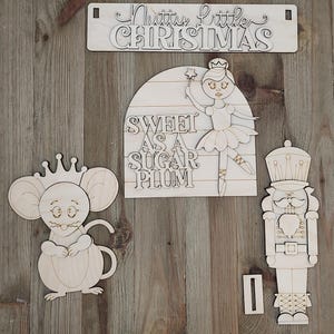 May include: A set of unfinished light-colored wooden cutouts featuring a rectangular sign with the text Nutty little CHRISTMAS, a semi-circular sign with the text SWEET AS A SUGAR PLUM, a mouse wearing a crown, and a nutcracker figure.