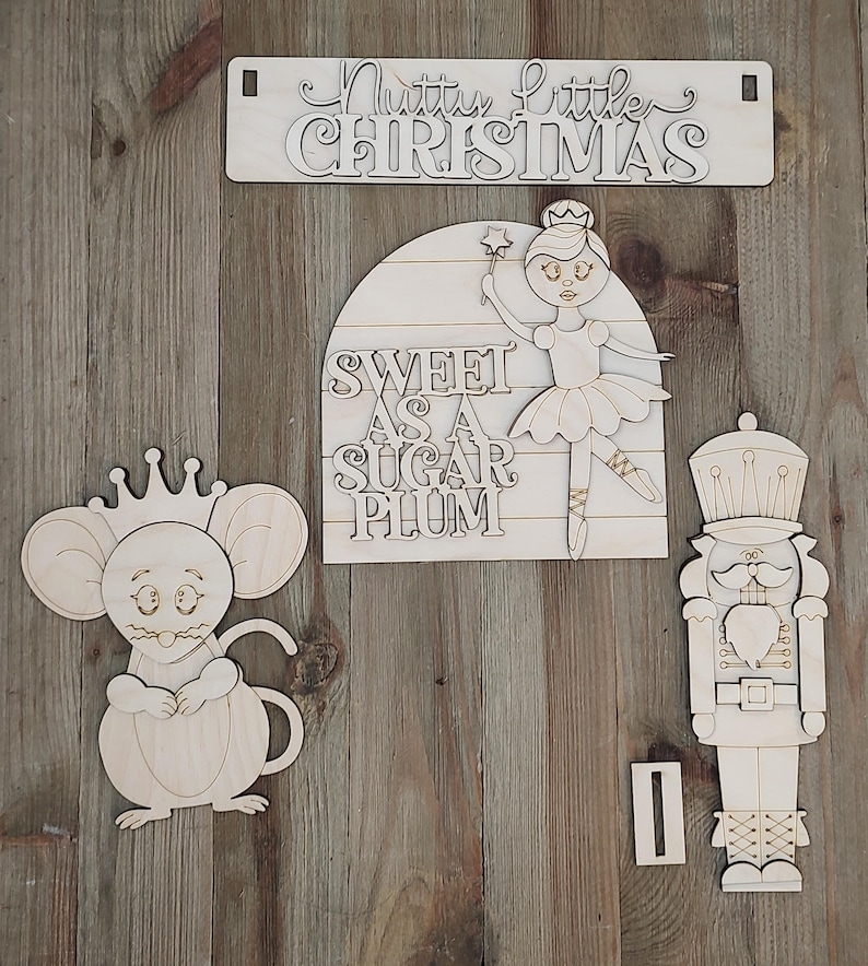 May include: A set of unfinished light-coloured wooden cutouts featuring a rectangular sign with the text Nutty little CHRISTMAS, a semi-circular sign with the text SWEET AS A SUGAR PLUM, a mouse wearing a crown, and a nutcracker figure.