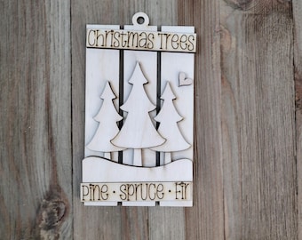 May include: A wooden Christmas ornament featuring three stylized Christmas trees. The ornament has the words "Christmas Trees" at the top and "Pine · Spruce · Fir" at the bottom. A small heart is cut out next to the trees.