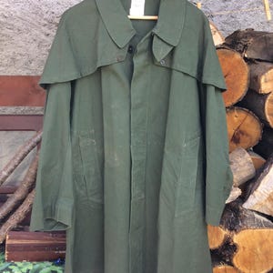 May include: An olive green vintage military coat with a cape-like collar and two front pockets. The coat is hanging on a wooden hanger. The coat is made of a durable fabric.