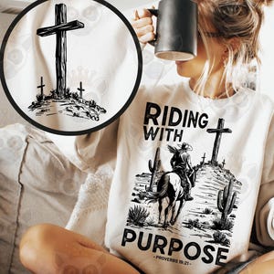 Könnte beinhalten: Ein weißes Sweatshirt mit einem schwarzen Aufdruck eines Cowboys auf einem Pferd, einem Kreuz und den Worten "RIDING WITH PURPOSE" und "PROVERBS 19:21". Das Sweatshirt hat auch eine runde Grafik eines Kreuzes.