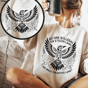 May include: Cream-colored sweatshirt with a black graphic of a bird with outstretched wings, surrounded by floral elements. The text "NO ONE IS ILLEGAL ON STOLEN LAND WE WALK ON NATIVE LAND" is printed below the bird. The image also includes a black mug.