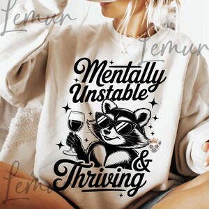 May include: Cream-colored sweatshirt with the text "Mentally Unstable & Thriving" in black script. A raccoon wearing sunglasses and holding a wine glass is featured in the center. The design includes star accents.